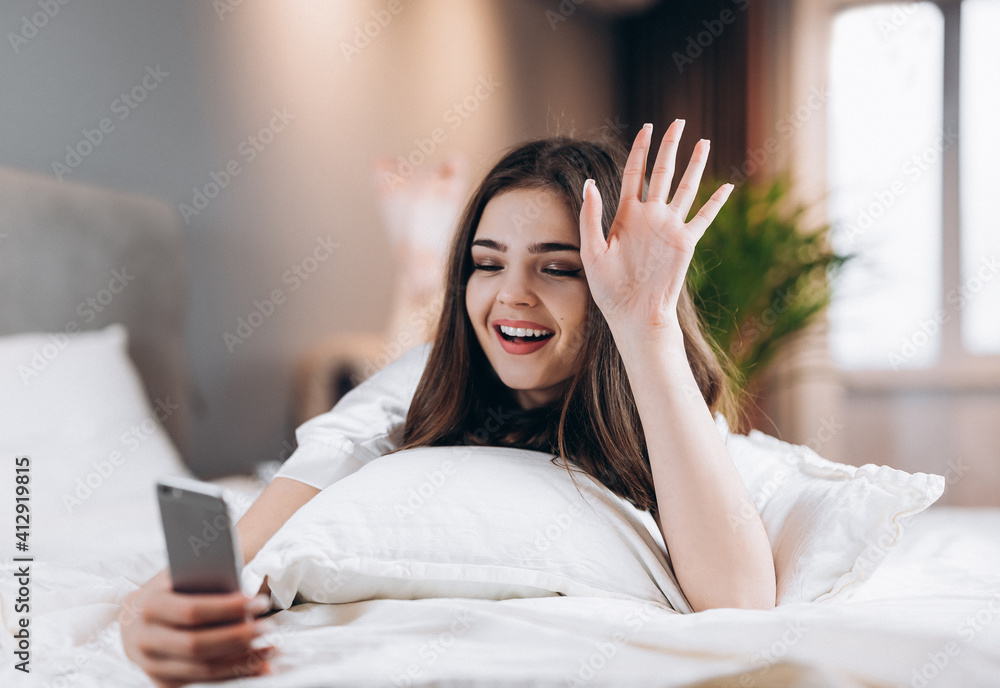 young woman looking at phone with a smile on her face. morning or evening in bed with a phone. Brunette scrolling the feed while lying in bed. Woman reading news on her phone or videocall