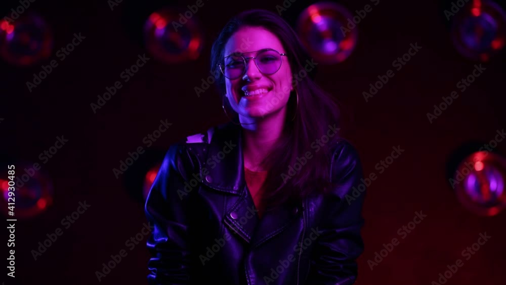 Portrait shot of the beautiful brunette girl in club atmosphere. Female in sunglasses standing in room in the pink and blue neon light, turning her head to the camera and laughing happily. Close up. Portrait shot of the beautiful brunette girl in club atmosphere. Female in sunglasses standing in room in the pink and blue neon light, turning her head to the camera and laughing happily. Close up.