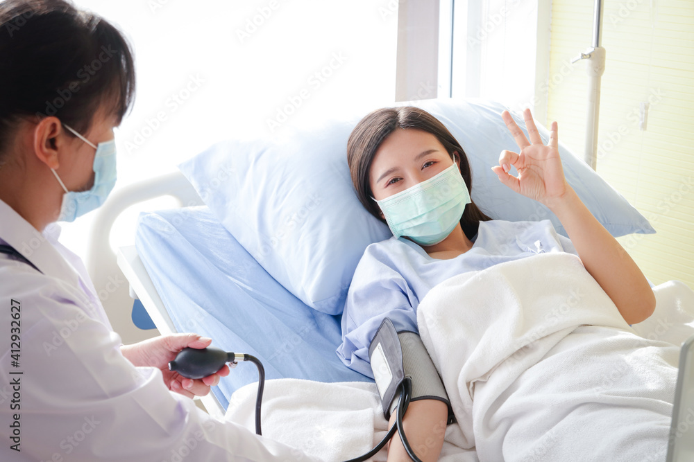 Asian female patients Wear a blue mask and lie on a hospital bed. Raise your hand. Okay. The doctor measures the patient's pressure. Concept of medical service, health insurance Asian female patients Wear a blue mask and lie on a hospital bed. Raise your hand. Okay. The doctor measures the patient's pressure. Concept of medical service, health insurance