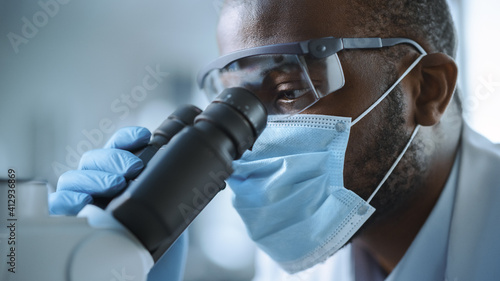 Close Up Shot of a Handsome Black Male Scientist Wearing Face Mask and Looking into the Microscope. Microbiologist Working on Molecule Samples in Modern Laboratory with Technological Equipment.