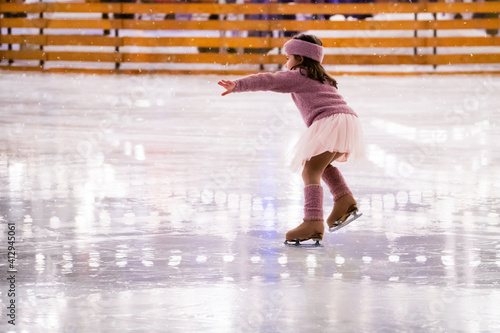 Fototapeta Naklejka Na Ścianę i Meble -  Little girl figure skater in pink sweater is skating on winter evening at an outdoor ice rink, back view