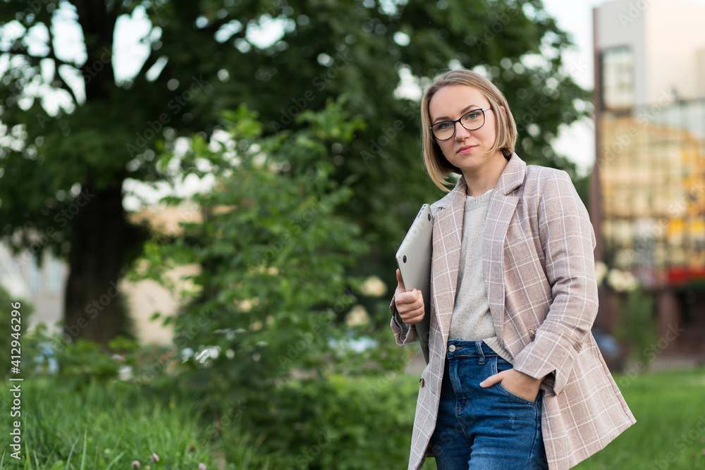 A girl of European appearance in glasses and a jacket with a laptop in the fresh air.An office-style woman stands in a park with her hand in her pocket. Remote work A girl of European appearance in glasses and a jacket with a laptop in the fresh air.An office-style woman stands in a park with her hand in her pocket. Remote work