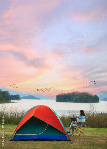 A sense of peace and serenity photo of female traveler sitting beside camping tent and using notebook laptop computer working from the lake side. There are group of birds flying in colorful sky