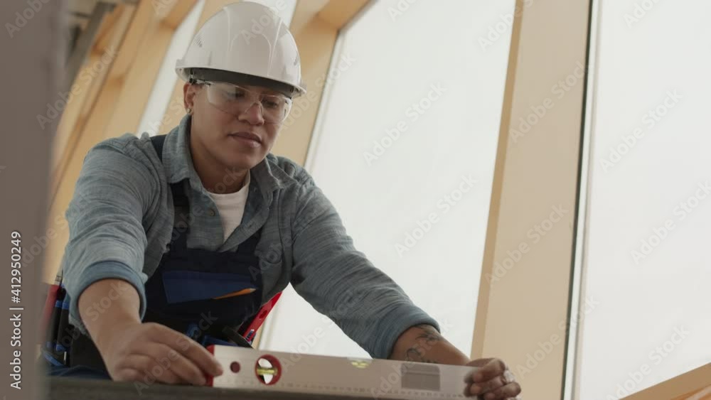 Low angle medium shot of professional young construction worker wearing helmet and goggles, sitting on metal structure of building under renovation, using bubble level in front of large window Low angle medium shot of professional young construction worker wearing helmet and goggles, sitting on metal structure of building under renovation, using bubble level in front of large window