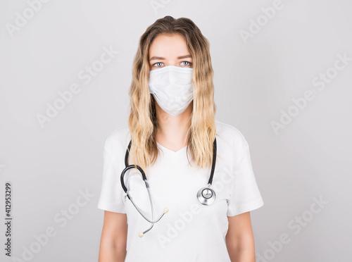 Medical concept of beautiful woman doctor in white coat with stethoscope, mask. Medical student. Woman hospital worker looking at camera and smiling, studio, gray background