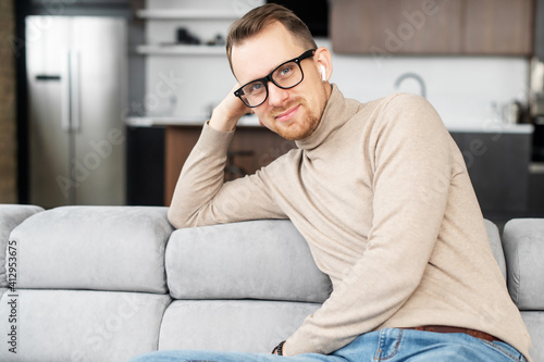 A portrait of handsome young man wearing stylish eyeglasses and smart casual wear, an attractive guy sitting on the comfortable couch at home, looks into the camera and smiles