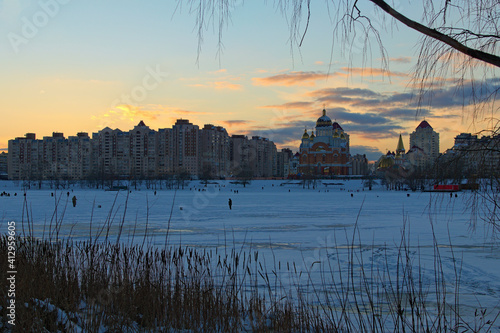 Scenic view of willow trees and poplars close to the frozen Dnieper River in Kyiv during sunset in winter. High buildings in the background. Obolon neighborhood. Kyiv, Ukraine