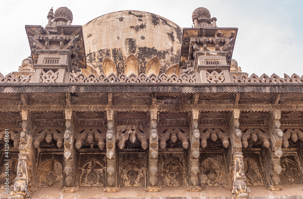 Vijayapura, Karnataka, India - November 8, 2013: Closeup of extensive brown stone sculptures under roof ledge with part of dome above at Ibrahim Rauza mausoleum under silver sky.