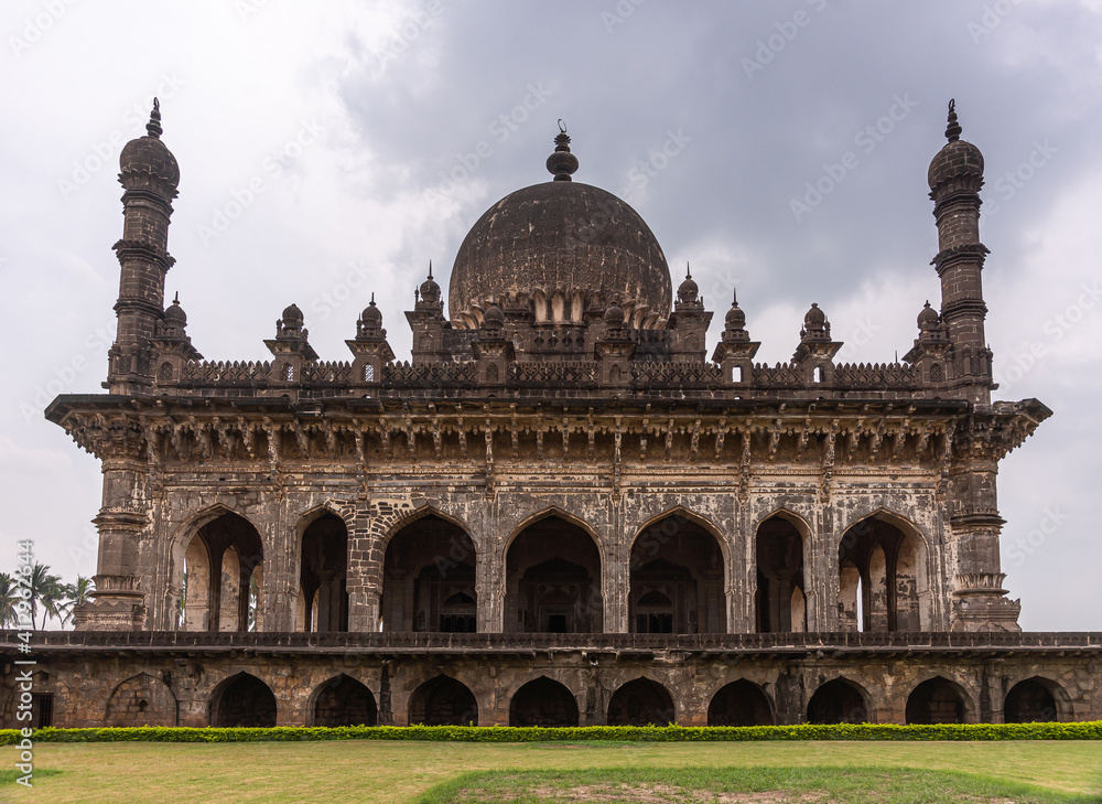 Fototapeta premium Vijayapura, Karnataka, India - November 8, 2013: Closeup of Ibrahim Rauza mausoleum. Brown stone under gray cloudscape with green lawn up front.