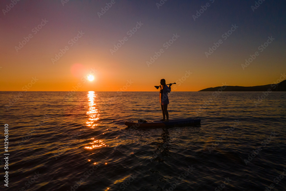 Young woman in two-piece swimsuit and shirt stands on SUP board with oar on her shoulders in the middle of sea against background of beautiful fiery sunset.