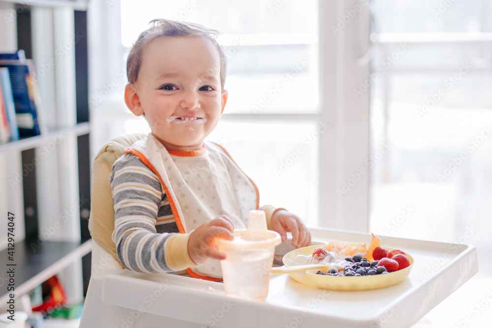 Cute Caucasian baby boy eating ripe berries and fruits with yogurt. Funny smiling child kid sitting in chair eating fresh berries and drinking water. Supplementary healthy food for toddler kids. Cute Caucasian baby boy eating ripe berries and fruits with yogurt. Funny smiling child kid sitting in chair eating fresh berries and drinking water. Supplementary healthy food for toddler kids.