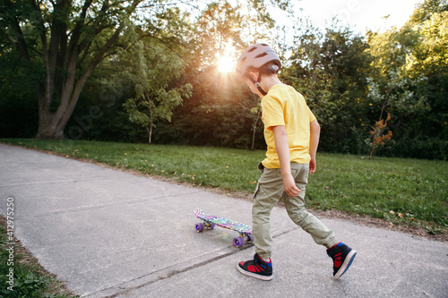 Boy in grey helmet riding skateboard on road in park on summer day. Seasonal outdoors children activity sport. Healthy childhood lifestyle. Boy learning to ride skateboard. View from back.