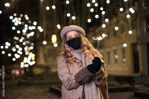 Young Woman wearing a Face Protective Mask Covid 19 holding a Sparkler and Making a Wish on New Year's Eve on a Cold Winter Evening, Outdoor with the City Lights in the Background, Copy Space, Holiday photo