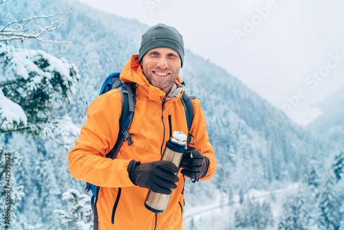 Laughing Man dressed bright orange softshell jacket with a hot drink thermos flask looking at camera while he trekking winter mountains route. Active people in the nature concept image. photo