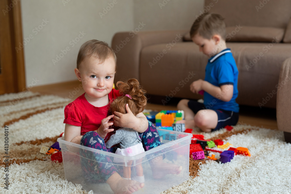 The older brother is playing on the carpet with the constructor, and the sister is sitting in a box from under the constructor. The older brother is playing on the carpet with the constructor, and the sister is sitting in a box from under the constructor.