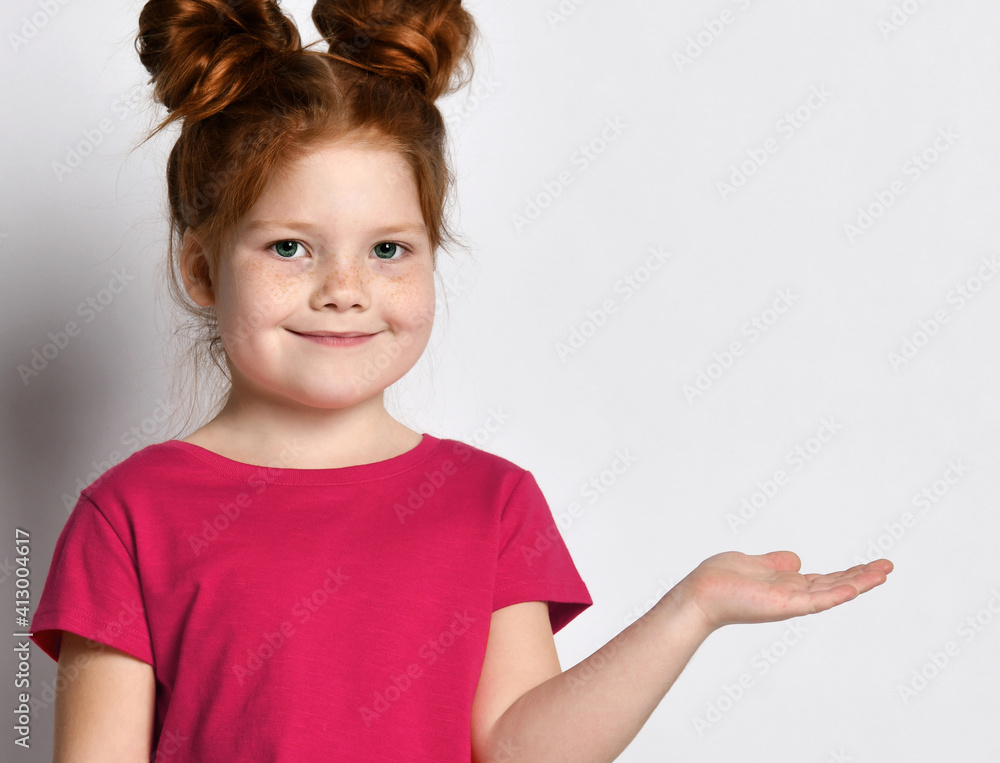 An intimate portrait of ginger Little cute girl with freckles on his face and a cute hairstyle hand points at the free space for text. Child posing on a gray background. An intimate portrait of ginger Little cute girl with freckles on his face and a cute hairstyle hand points at the free space for text. Child posing on a gray background.