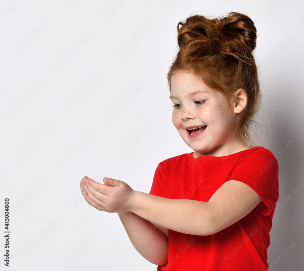 Close-up of a pleasantly surprised red-haired girl looking happily at something in her palms. The girl stands on a gray background near the place for the text. The concept of positive children Close-up of a pleasantly surprised red-haired girl looking happily at something in her palms. The girl stands on a gray background near the place for the text. The concept of positive children