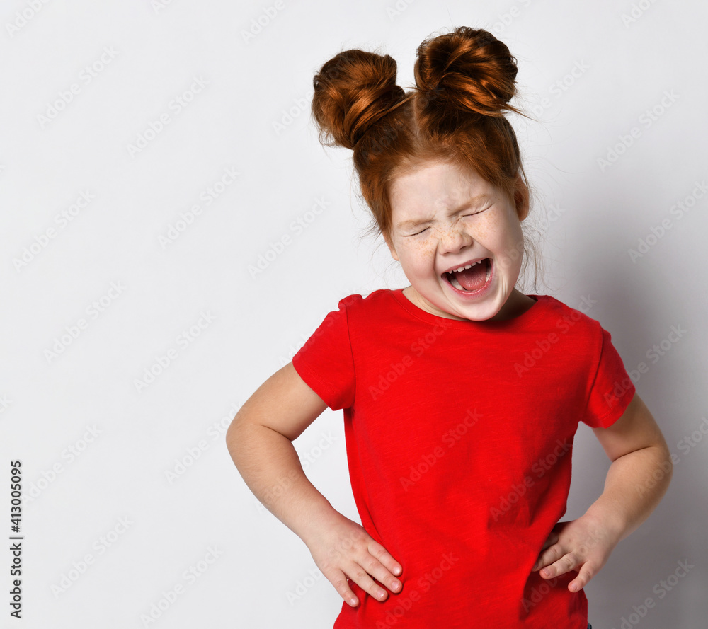Little girl with red hair and freckles holds her hands on her waist and screams loudly. Child in a red T-shirt posing on a background of a gray wall in the studio. Concept of children's emotions. Little girl with red hair and freckles holds her hands on her waist and screams loudly. Child in a red T-shirt posing on a background of a gray wall in the studio. Concept of children's emotions.