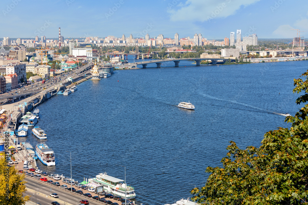 A landscape of summer Kyiv with a view of the Dnipro embankment in the old Podil district, a river station, piers, river trams and pleasure boats.