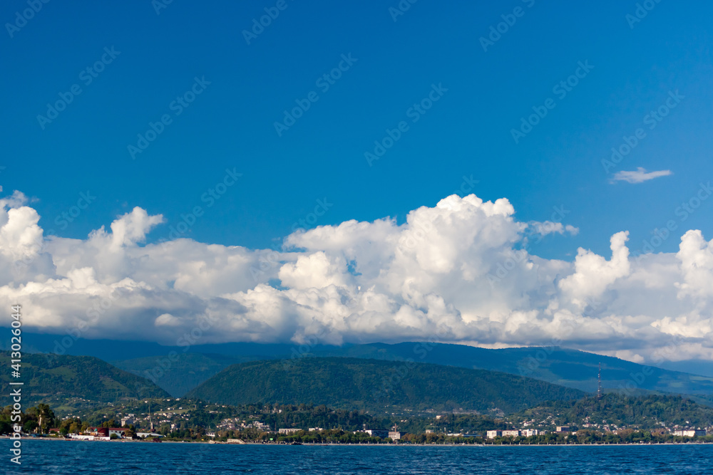Sea and mountains against the blue sky with white clouds, at the bottom of the mountains are buildings Sea and mountains against the blue sky with white clouds, at the bottom of the mountains are buildings