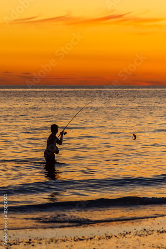 Silhouette of a boy standing in the ocean catching a fish as the sun sets in the background and creates an orange glow in the sky