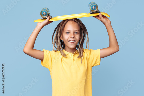 Positive little boy with african dreads holding skateboard on the head over blue background.
