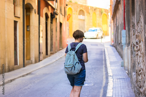 A tourist girl with a striped backpack walks down a sun-drenched European street. No face visible. Traveling through Spain, Barcelona.