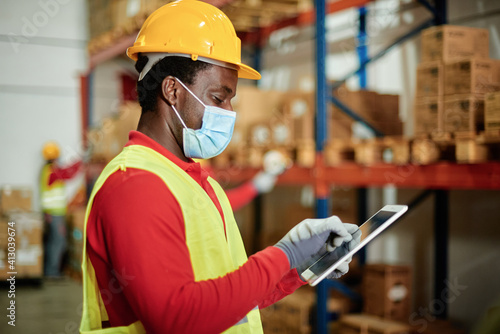 Afro male worker using a tablet checking the transport of a box in a warehouse wearing a safety mask and work waistcoat.