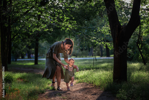 The baby girl takes the first steps, a young mother holding a child by the hands, insuring, helping to walk along the path of the park amid green trees and grass. Parental care, walks in the fresh air