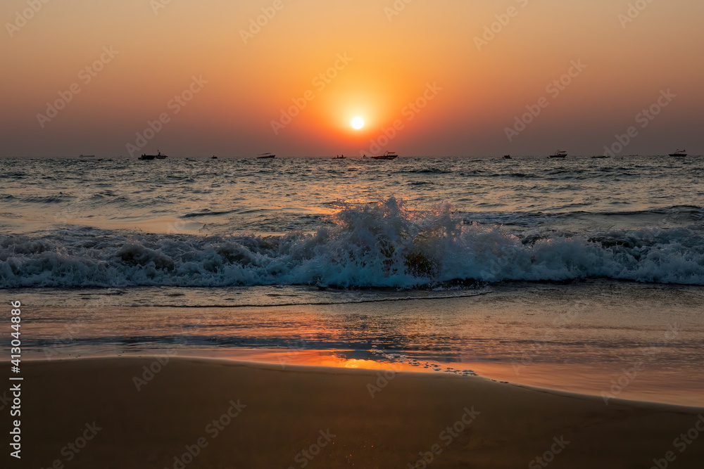 Scenic Sunset on beach in South Asia, white wave hitting sandy coastline. A lot of fishing boats on horizon line in Indian Ocean. Clear evening sky beautifully highlighted by orange red Sun