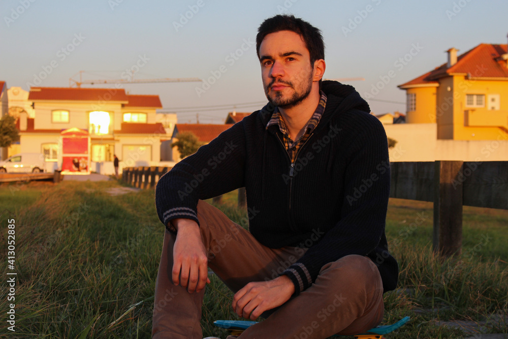 Young man posing with his skateboard outdoor, staring into the distance, great sunset light. Gay sitting on his skateboard in the grass. Young man posing with his skateboard outdoor, staring into the distance, great sunset light. Gay sitting on his skateboard in the grass.