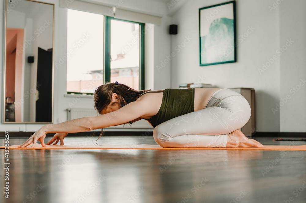 Young woman in white scribbled leggings doing active child's pose yoga posture with fingertips on the floor in an empty yoga studio. Concept: wellness, self care during pandemic, active mobility.
