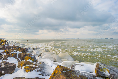 Bild auf Leinwand Snowy iced rocks of a dike along a white frozen lake in wetland under a blue clo