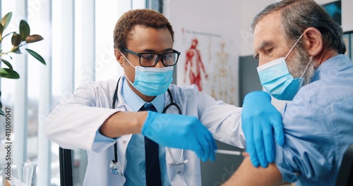 Close up portrait of cheerful young African American handsome professional man doctor in white coat wearing medical mask making coronavirus vaccine injection to Caucasian old patient in clinic