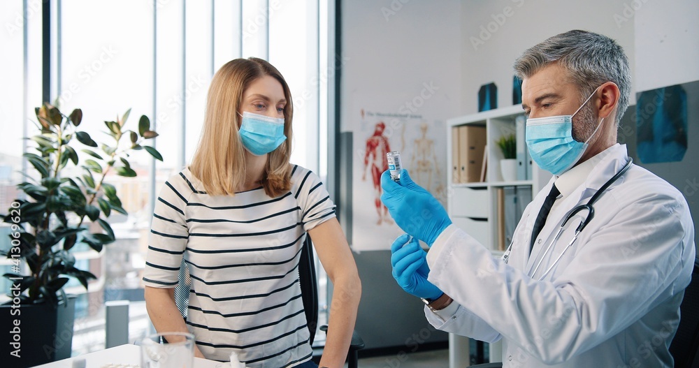 Fototapeta premium Portrait of professional handsome middle-aged doctor virologist in medical mask and white coat injecting coronavirus vaccine to Caucasian happy adult woman patient sitting in hospital, healthcare