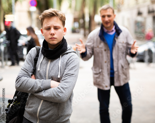 Portrait of troubled teen boy standing with arms crosse outdoors while man stranger scolding him