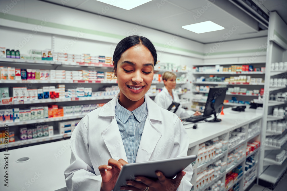Successful young female pharmacist using digital tablet in drugstore with colleague using computer in background