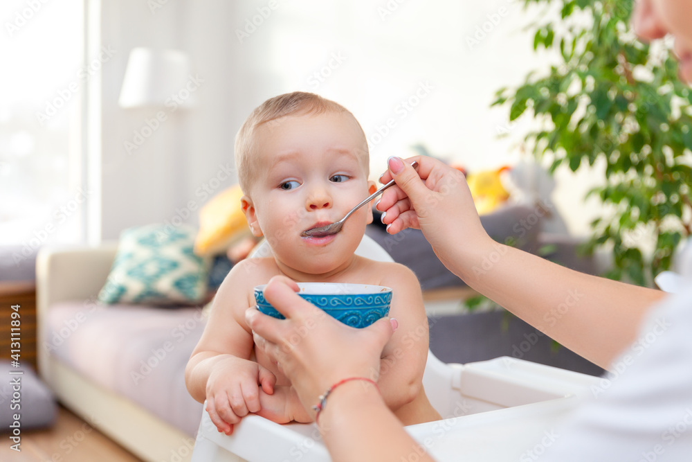Mother's hand feeds funny cute happy blond baby toddler with spoon Mother's hand feeds funny cute happy blond baby toddler with spoon