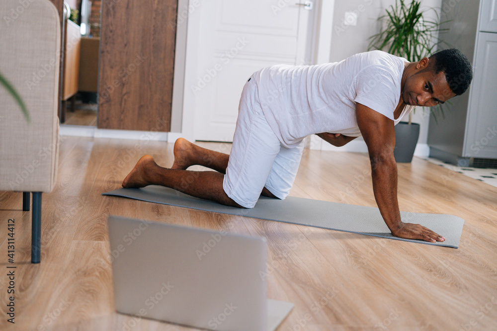 Muscular African-American fitness instructor speaking by laptop webcam streaming online training exercise classes sitting on floor. Concept of sport training at home gym.