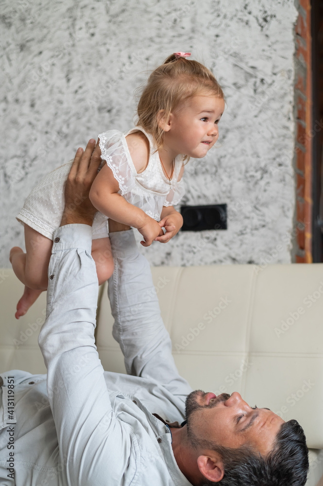 Naklejka premium A Caucasian man lies on the couch and holds a little daughter above him. Caring loving dad raised the child up. Happy fatherhood.