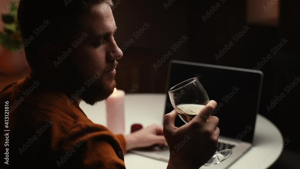 Close-up back view of thoughtful young man drinking alcohol from glass during working on laptop at nighttime. Bearded male sitting at home and drinking wine while browsing internet on computer. Close-up back view of thoughtful young man drinking alcohol from glass during working on laptop at nighttime. Bearded male sitting at home and drinking wine while browsing internet on computer.