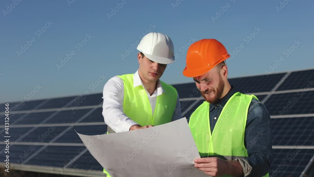Two engineers are looking at the project blueprints. They are discussing all the stages. Solar panels in the background. 4K.