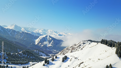 View from the height of the snowy mountains and forest. Huge clouds cover the hill in the distance. People are visible in places. Someone is running, resting, having a picnic or climbing to the top.