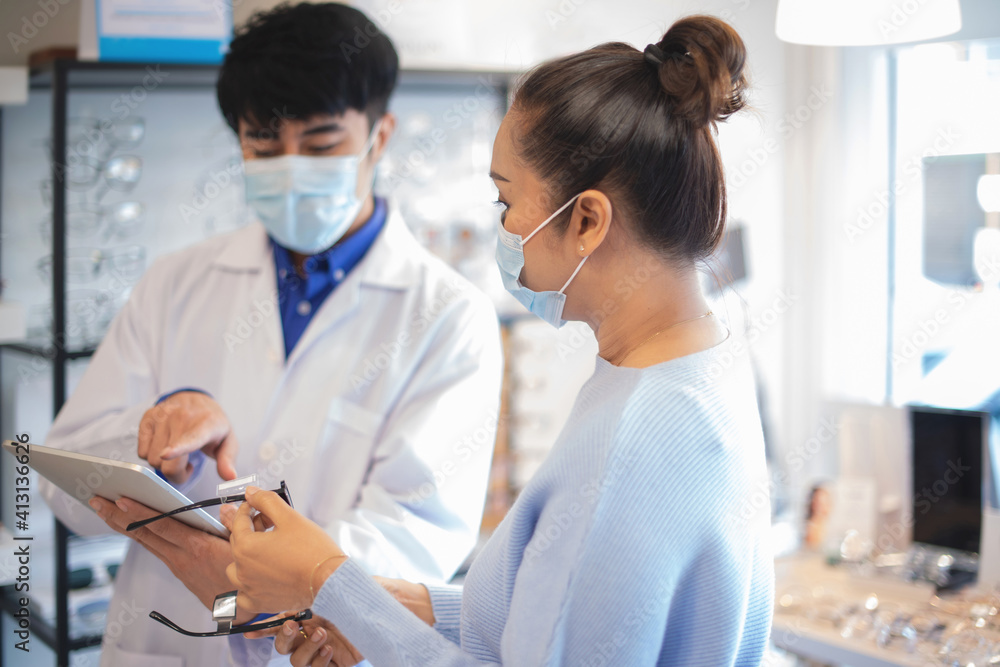 Asian optometrist in white coat and medical mask gives senior woman advice on new model of glasses Asian optometrist in white coat and medical mask gives senior woman advice on new model of glasses