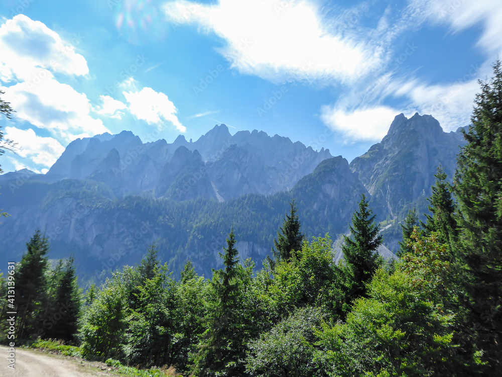 Gravelled road in the mountains with the view on high Alps in the region of Gosau, Austria. The lower mountains are overgrown with dense forest. The chains in the back are stony and barren. Sunny day