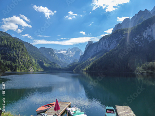 Panoramic view on Gosau lake, with Dachstein glacier in the back in Austrian Alps. The lake is surrounded by high mountains, overgrown with tall trees. Few colorful boats anchored to a pier. Serenity