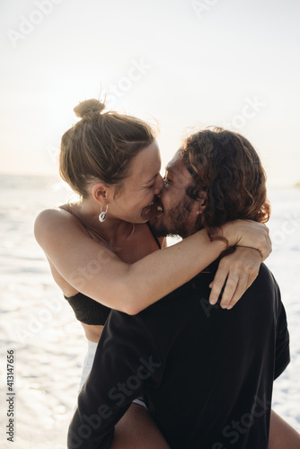The man raised his beloved in his arms while she wrapped her arms around his neck laughing and kissing him against the background of the ocean and sand. High quality photo