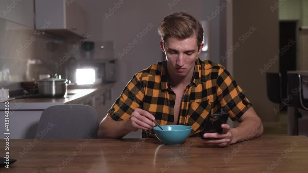a Caucasian man of appearance sits at a table, eats with a spoon and reads the news on his phone. Bachelor breakfast concept a Caucasian man of appearance sits at a table, eats with a spoon and reads the news on his phone. Bachelor breakfast concept