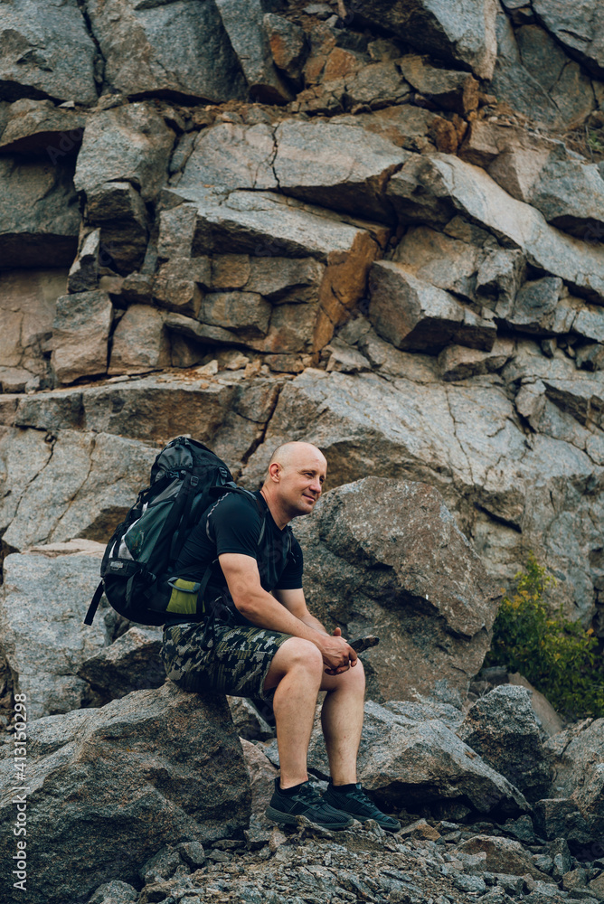 Traveler with a backpack sits on cliff in mountains. View from a side on a man surrounded by big rocks. Hiking in mountains.