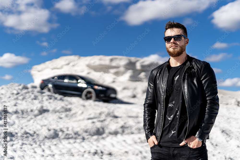 Cute stylish fashion model guy on photosession in a quarry. Luxury car on the background. rich man in leather jacket and sunglasses. Cute stylish fashion model guy on photosession in a quarry. Luxury car on the background. rich man in leather jacket and sunglasses.